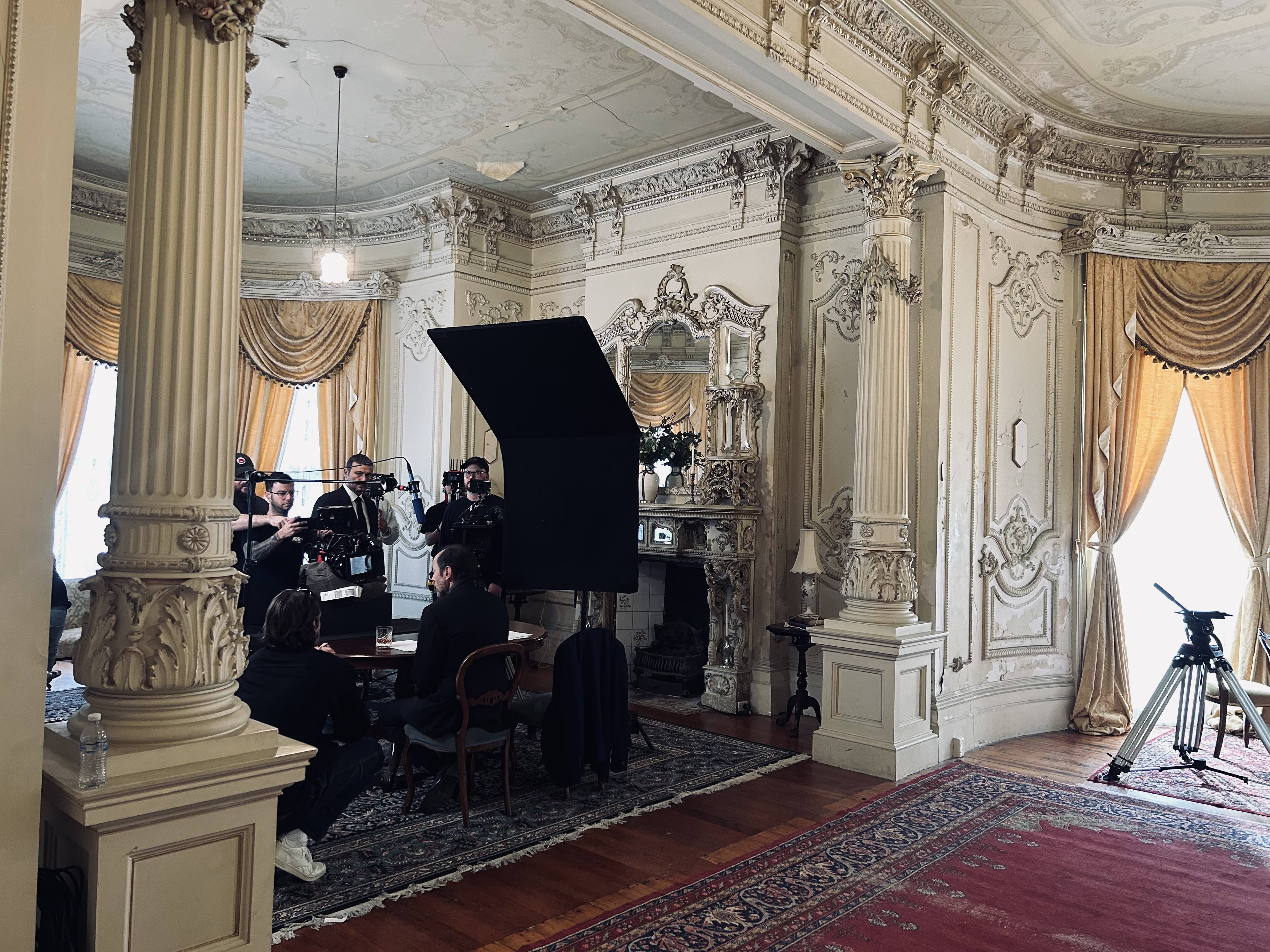 Filming crew in an ornate classical ballroom with carved columns, gold drapes, and a large black monitor.