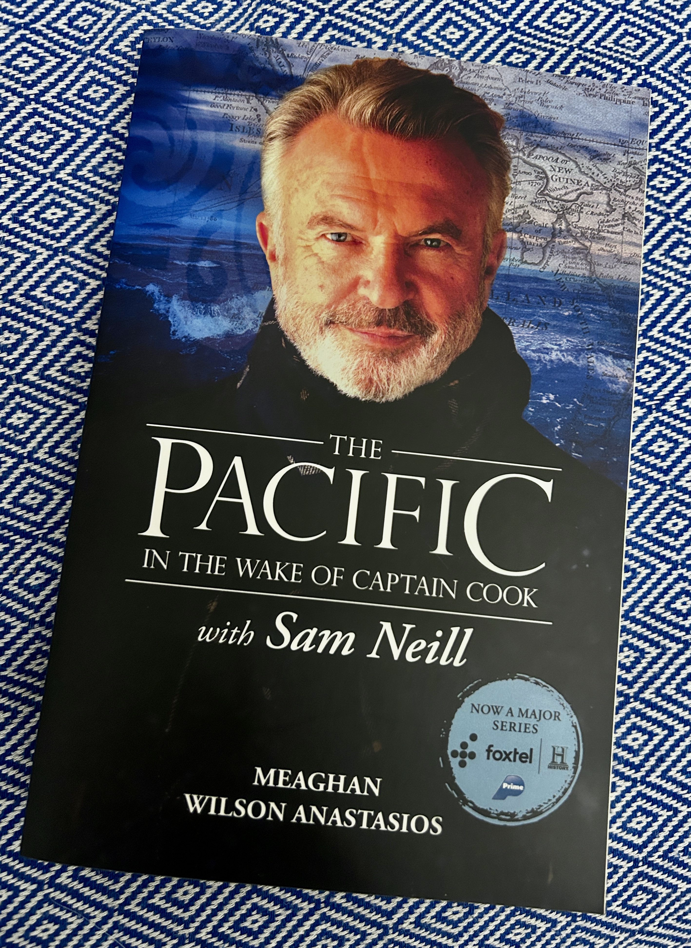 Book cover showing a close-up of a man with gray hair and beard, against a map and ocean background; title reads The Pacific: In the Wake of Captain Cook, with Sam Neill (text visible).